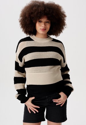 Chunky Strickpullover mit schwarz-beigem Streifenmuster, geripptem Rundhalsausschnitt, weiten Ärmeln und strukturiertem Stoff, kombiniert mit schwarzen Shorts.