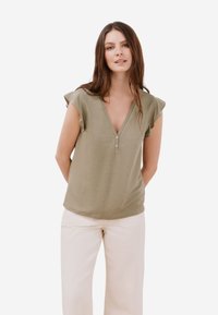 Olivgrön t-shirt med kort ärm och V-ringning samt två knappar framtill. Mjuk tyg med en avslappnad passform och volangkanter på ärmarna.