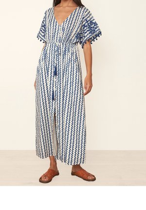 Frau trägt ein blau-weißes Maxikleid mit Zickzackmuster, kurzen Ärmeln und braunen Ledersandalen und steht auf einem hellen Holzboden.