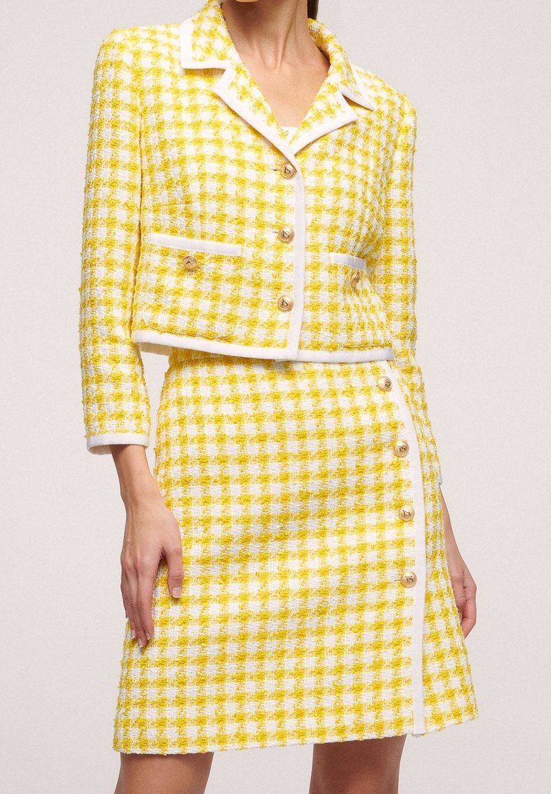 Žlutý tvídový blejzr a sukně se vzorem kohoutí stopy, bílým lemem, zlatými knoflíky a zkráceným sáčkem s dvěma předními kapsami.