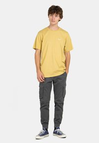 Camiseta de algodón amarilla de manga corta con un pequeño logo blanco en el pecho, combinada con pantalones cargo grises y zapatillas azules.