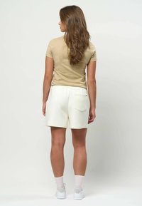 Femeie cu păr lung și castaniu, văzută de spate, purtând un top bej cu mâneci scurte, pantaloni scurți albi cu buzunar pe spate, șosete albe și adidași albi.