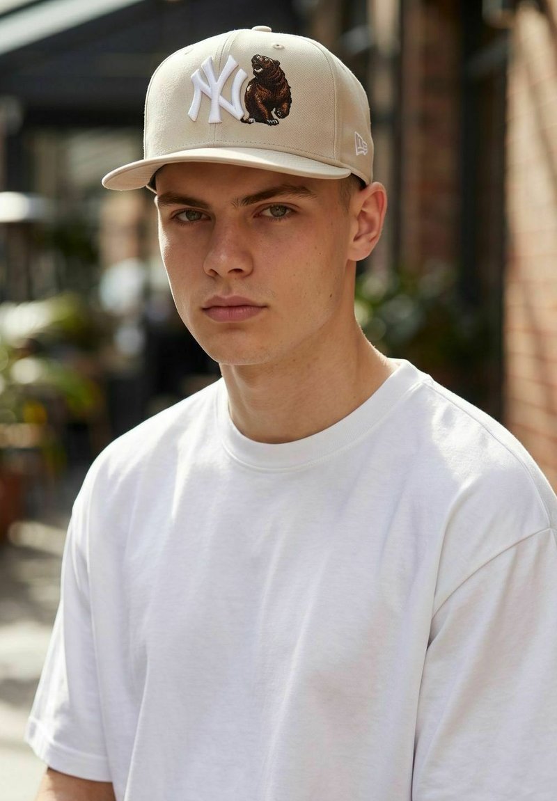 Young man with a neutral expression wearing a beige cap with white NY letters and a small bear emblem, and a plain white t-shirt.