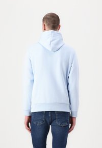 Sudadera azul claro con capucha ajustable, puños y dobladillo acanalados. La tela parece suave y texturizada. Llevada con jeans oscuros.
