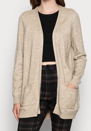 Woman wearing an open beige knit cardigan with front pockets, a black crop top, and black plaid pants. One hand in pocket.