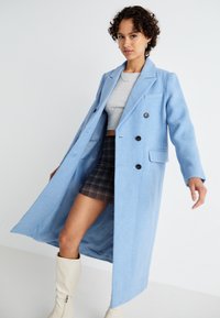 Casaco azul claro, de duplo botão, com lapelas recortadas, dois bolsos frontais e botões pretos; combinado com um top cinza e uma saia mini de padrão xadrez.