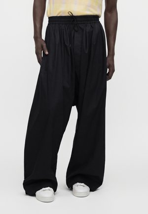 Personne portant un pantalon noir ample avec une taille élastique et des baskets blanches, debout devant un fond clair uni.