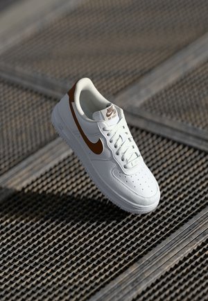 Biały sneaker Nike Air Force 1 z brązowym logo i akcentami, wyświetlony na teksturowanej metalowej powierzchni kratowej.