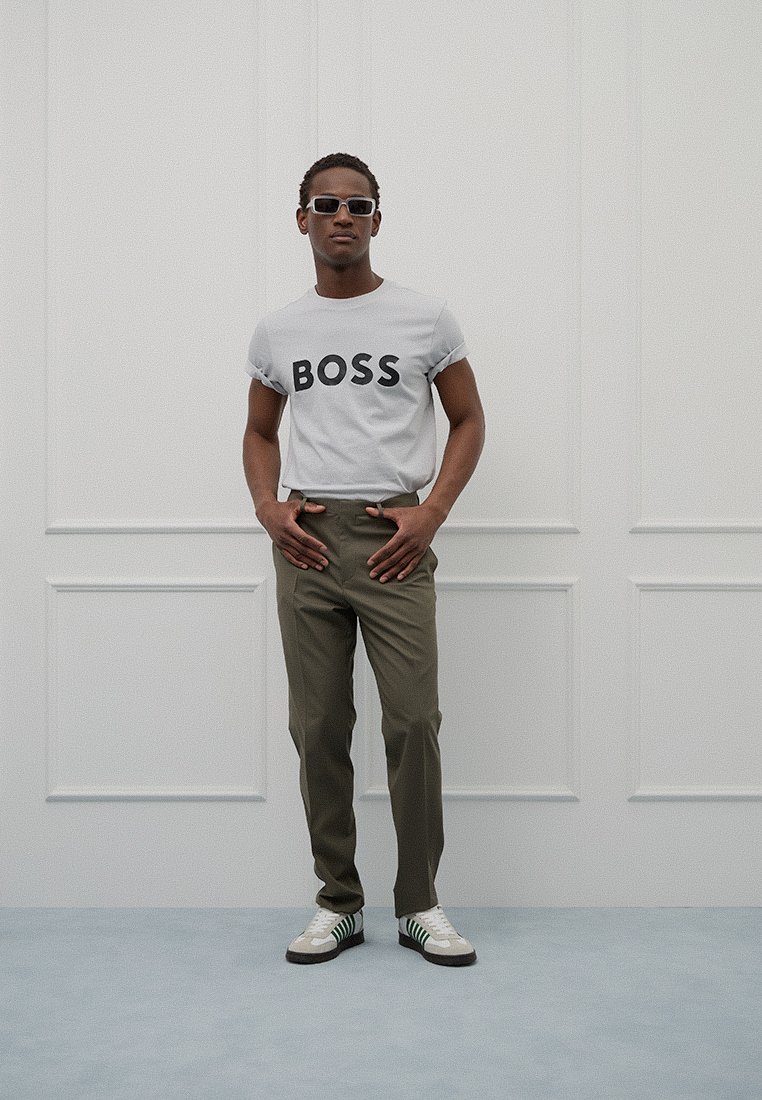 Camiseta gris con la palabra "BOSS" impresa en negro, pantalones oliva, zapatillas blancas con rayas verdes y gafas de sol. Diseño moderno y sencillo.