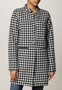 Svart och vit hounds-tooth-mönstrad kappa med kragdesign, två framfickor och knappstängning, visad på en modell.
