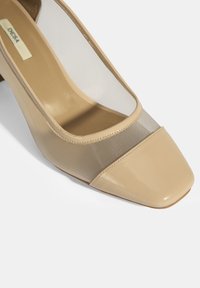 Pompe en cuir verni beige avec un panneau en maille au niveau des orteils, de forme carrée, talon bas et texture lisse. Aucuns motifs ni accessoires notables présents.