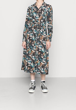 Frau trägt ein langärmeliges, knielanges Blumenmusterkleid mit Kragen in den Farben Schwarz, Weiß, Türkis und Beige, kombiniert mit schwarzen High-Top-Sneakern und weißen Socken.