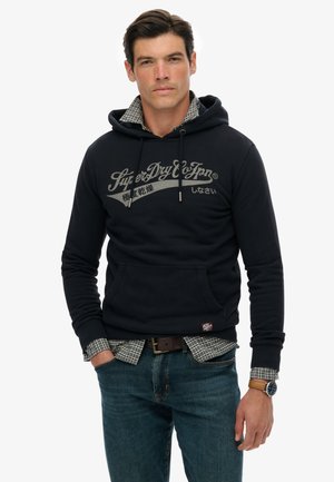 Marineblå hoodie med forlommer, med lys grå branding. Bæres over en ternet skjorte, sammen med blå jeans og et brunt bælte.