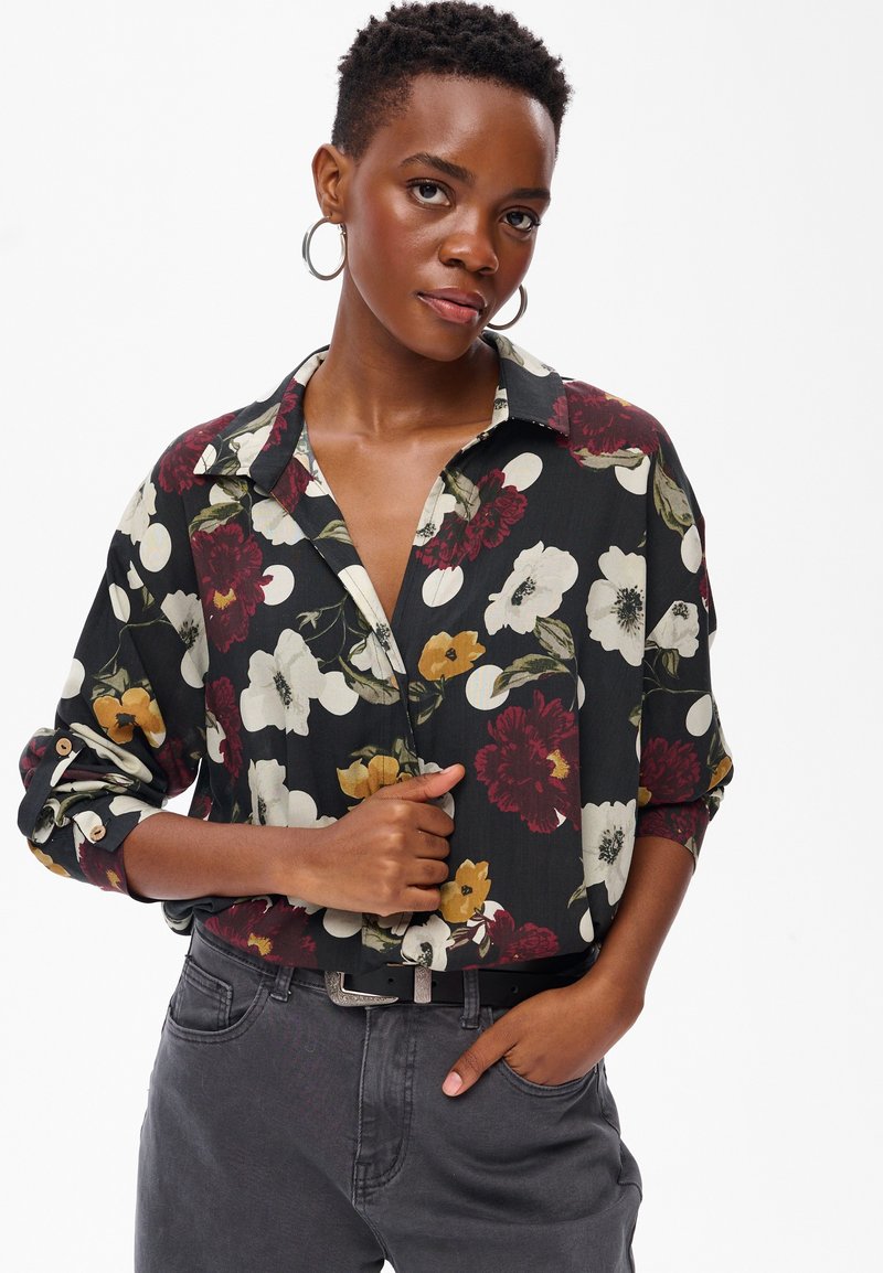 Jeune femme aux cheveux courts portant des boucles d'oreilles en anneau, une chemise noire à fleurs avec des manches retroussées, et un jean gris foncé avec une ceinture noire.