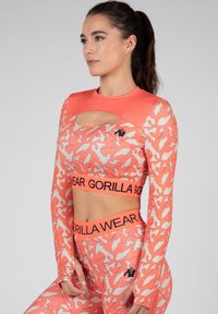 Koraalkleurige crop top met lange mouwen en uitsnede, met een beige abstract patroon en zwart logo. Gecombineerd met bijpassende leggings en een geprint tailleband.