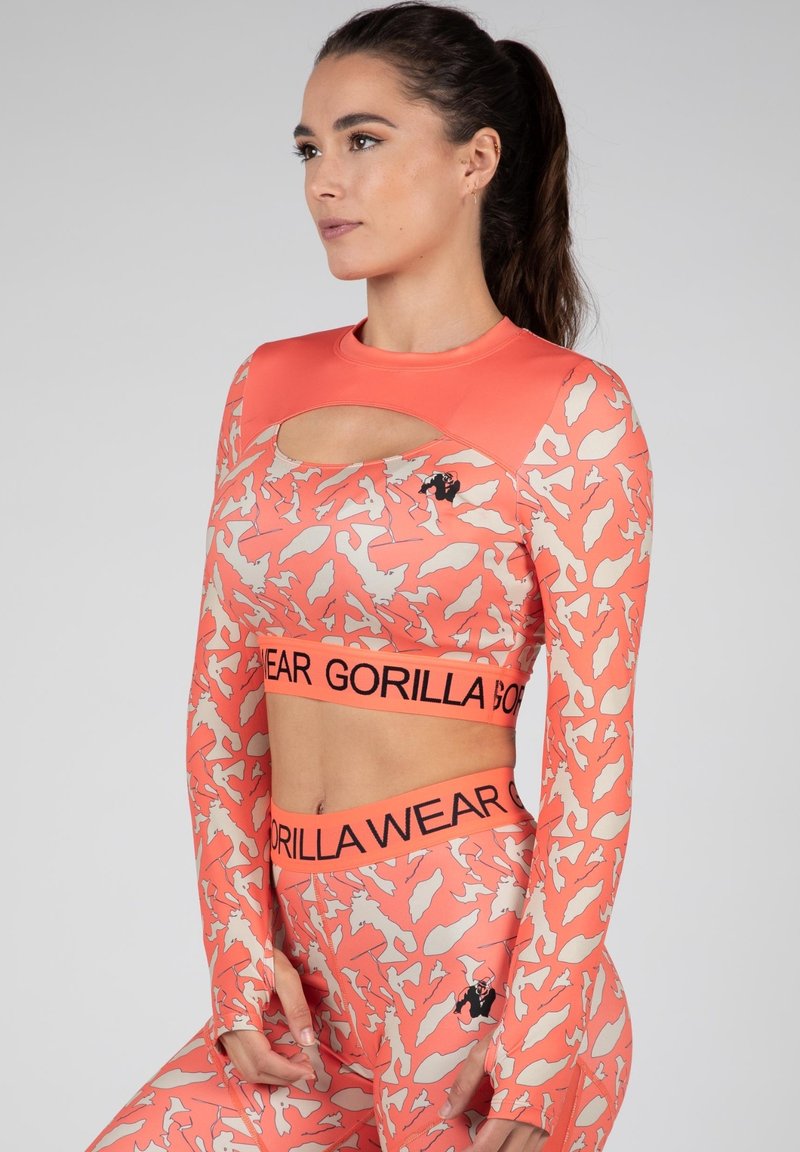 Koraalkleurige crop top met lange mouwen en uitsnede, met een beige abstract patroon en zwart logo. Gecombineerd met bijpassende leggings en een geprint tailleband.