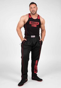 Zwarte tanktop en sweatsbroek met rode accenten. Tekst "GORILLA WEAR" op de voorkant en zijkant. Sportieve pasvorm met afgeknotte mouwen, die spieren benadrukken.