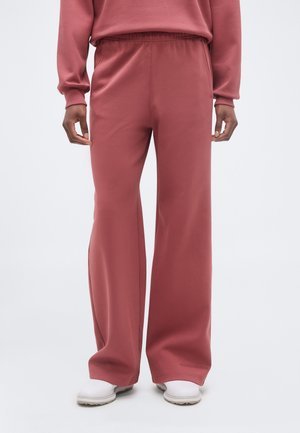 Inimene kannab lahtisi sobivaid tuhmi roosavaid dressipükse elastse taljega ja valgeid kingi ühtlase hele tausta ees.