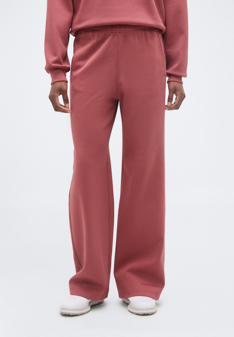 Άτομο που φοράει φαρδιές, σάπιες ρόζ φόρμες με ελαστική μέση και λευκά παπούτσια σε απλό ανοιχτό φόντο.