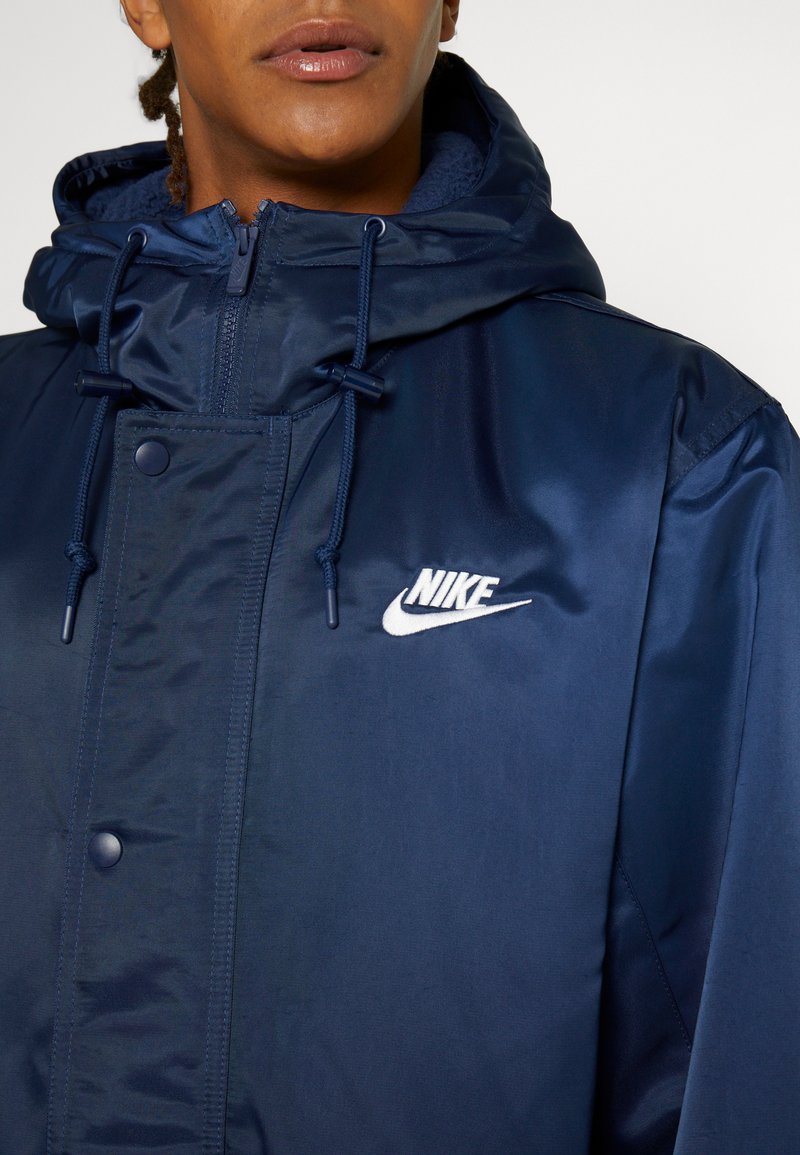 Granatowa kurtka Nike z błyszczącego materiału, regulowany kaptur ze sznurkiem oraz małe białe logo na lewej piersi.