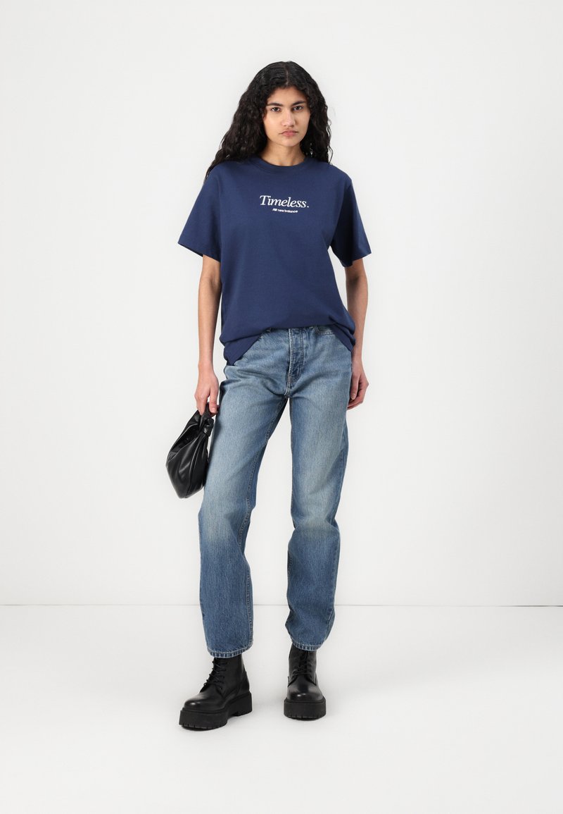 T-Shirt en coton bleu marine avec "Timeless." imprimé en blanc, associé à un jean bleu délavé et des bottes noires à semelle épaisse. Le modèle tient un sac noir.