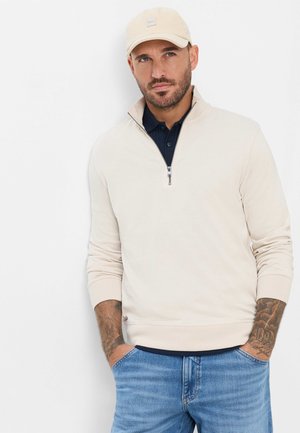 Mann mit Bart und Tattoos, der einen beigen Sweatshirt mit Reißverschluss, blaue Jeans, eine cremefarbene Mütze und darunter ein dunkles Hemd trägt, steht mit den Händen in den Taschen.