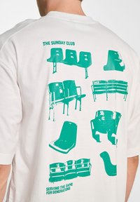 White t-shirt featuring green graphics of various chair designs and the text "THE SUNDAY CLUB" at the top and "SERVING THE GAME FOR GENERATIONS" at the bottom.
