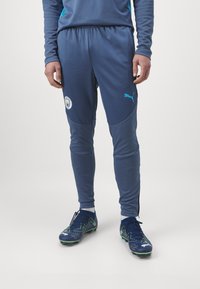 Pantaloni da allenamento Puma blu, con cintura in vita. Pannelli laterali e dettaglio del logo. Indossati con scarpini da calcio blu con accenti verdi.