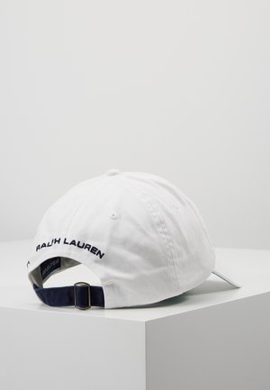Biała czapka bejsbolowa Ralph Lauren z regulowanym granatowym paskiem i wyhaftowaną nazwą marki z tyłu, spoczywająca na białej powierzchni.