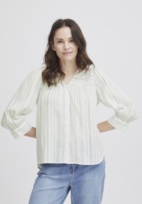 Camicia a righe chiare con bottoni in tessuto morbido, con scollatura a V e maniche a tre quarti, dalla vestibilità comoda e con fessure laterali. Toni di blu e verde.