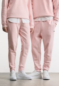 Δύο άτομα φορούν ταιριαστά ανοιχτό ροζ φούτερ και φόρμες, λευκά μπλουζάκια από μέσα και λευκά αθλητικά παπούτσια, στέκονται δίπλα-δίπλα.