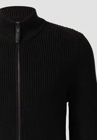 Schwarzer gestrickter Reißverschlusspullover mit hohem Kragen, geripptem Muster und metallicfarbener Reißverschluss, der eine taillierte Passform zeigt.