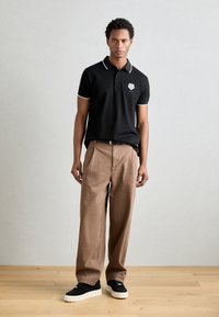 Zwarte polo met witte accenten en geborduurd logo, gecombineerd met losse beige broek en zwart sneakers met witte zolen.
