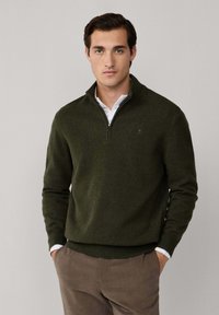Suéter verde oliva con cremallera, con puños y dobladillo acanalados, hecho de material suave, superpuesto sobre una camisa blanca con cuello, de tela lisa.