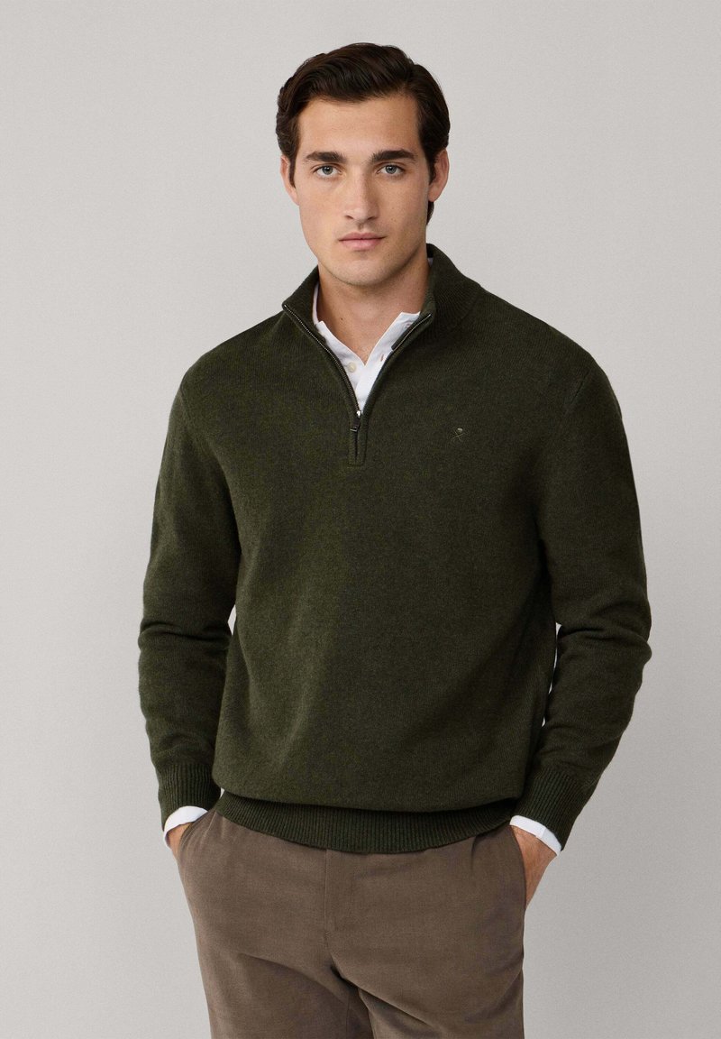 Suéter verde oliva con cremallera, con puños y dobladillo acanalados, hecho de material suave, superpuesto sobre una camisa blanca con cuello, de tela lisa.