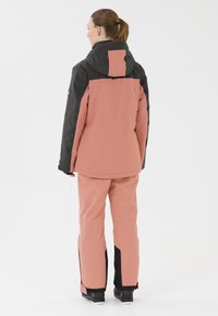 Rosa und schwarze Skijacke mit Kapuze, langen Ärmeln und passender roter Hose. Das Material scheint wasserdicht zu sein, mit glatter Oberfläche und minimalen Akzenten.