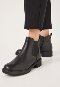 Stivali neri in pelle fino alla caviglia con inserti elastici laterali, superficie liscia, tacco a blocco e dettaglio con zip sul retro. La suola presenta scanalature antiscivolo.