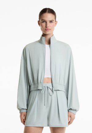 Jachetă cropped de culoare verde deschis, fabricată dintr-un material moale, cu un guler înalt, bandă elastică la talie și mâneci relaxate, asortată cu pantaloni scurți corespunzători.
