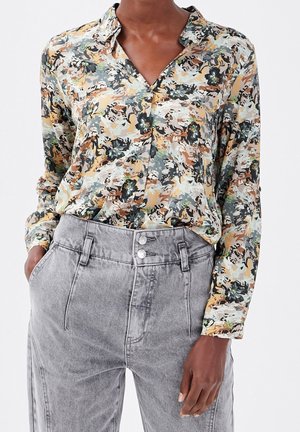 Personne portant une chemise à manches longues fleurie, boutonnée, rentrée dans un jean gris taille haute avec les mains dans les poches.