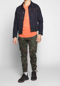 Czarna kurtka dżinsowa, pomarańczowa koszulka i zielone bojówki moro. Buty czarne z grubą podeszwą. Ręce trzymane luźno po bokach z lekkim zaciskiem.