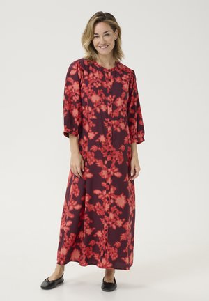 Lång, burgundy klänning med ett blommigt röd mönster, knäppning fram och ¾ ärmar. Lättviktsmaterial, flödande silhuett, bärs med svarta ballerinaskor.