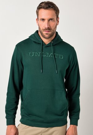 Uomo che indossa una felpa verde scuro con "UNLMTD" ricamato sul petto e pantaloni beige, in piedi contro uno sfondo chiaro e uniforme.