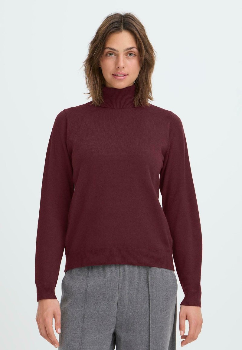 Burgundy polokrage tröja i mjukt stickat tyg med långa ärmar; har en figurnära silhuett och ribbade mudd och fåll. Bärs med grå byxor.