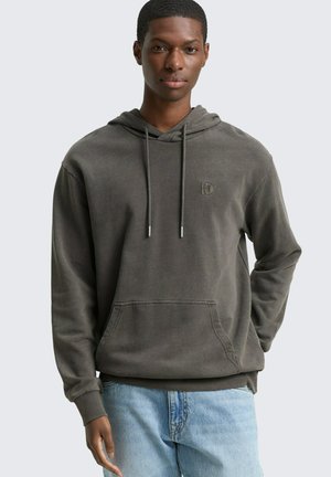 Młody mężczyzna w wyblakłej ciemnoszarej bluzie z kapturem i kieszenią z przodu oraz jasnoniebieskich dżinsach, stojący na neutralnym tle.