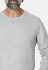 Hombre con barba que lleva un suéter de punto texturizado gris claro de mangas largas frente a un fondo simple.