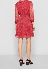 Mujer vistiendo un vestido rojo floral de longitud a la rodilla con mangas largas transparentes y sandalias de tacón bloque negro, de pie contra un fondo blanco liso.