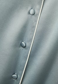 Satin shirt i blå-grå med en mjuk yta, med runda matchande knappar och kontrasterande vit piping längs knappslån.