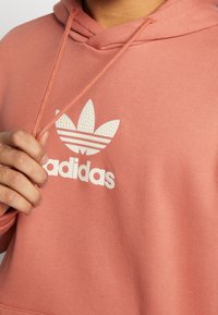 Rosa Adidas-huvtröja med en strukturerad vit logotyp, dragsko på huvan och kängurufickor. Mjuk tygkvalitet med ribbade manschetter och nederkant.