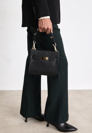 Personne portant un pantalon vert foncé et des talons aiguilles noirs tient un petit sac à main noir avec des accents dorés sur un fond uni.