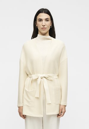 Kobieta z długimi ciemnymi włosami, ubrana w jasnobeżowy prążkowany kardigan przewiązany w talii, na wysokim golfie i dopasowanych spodniach, na jednolitym tle.